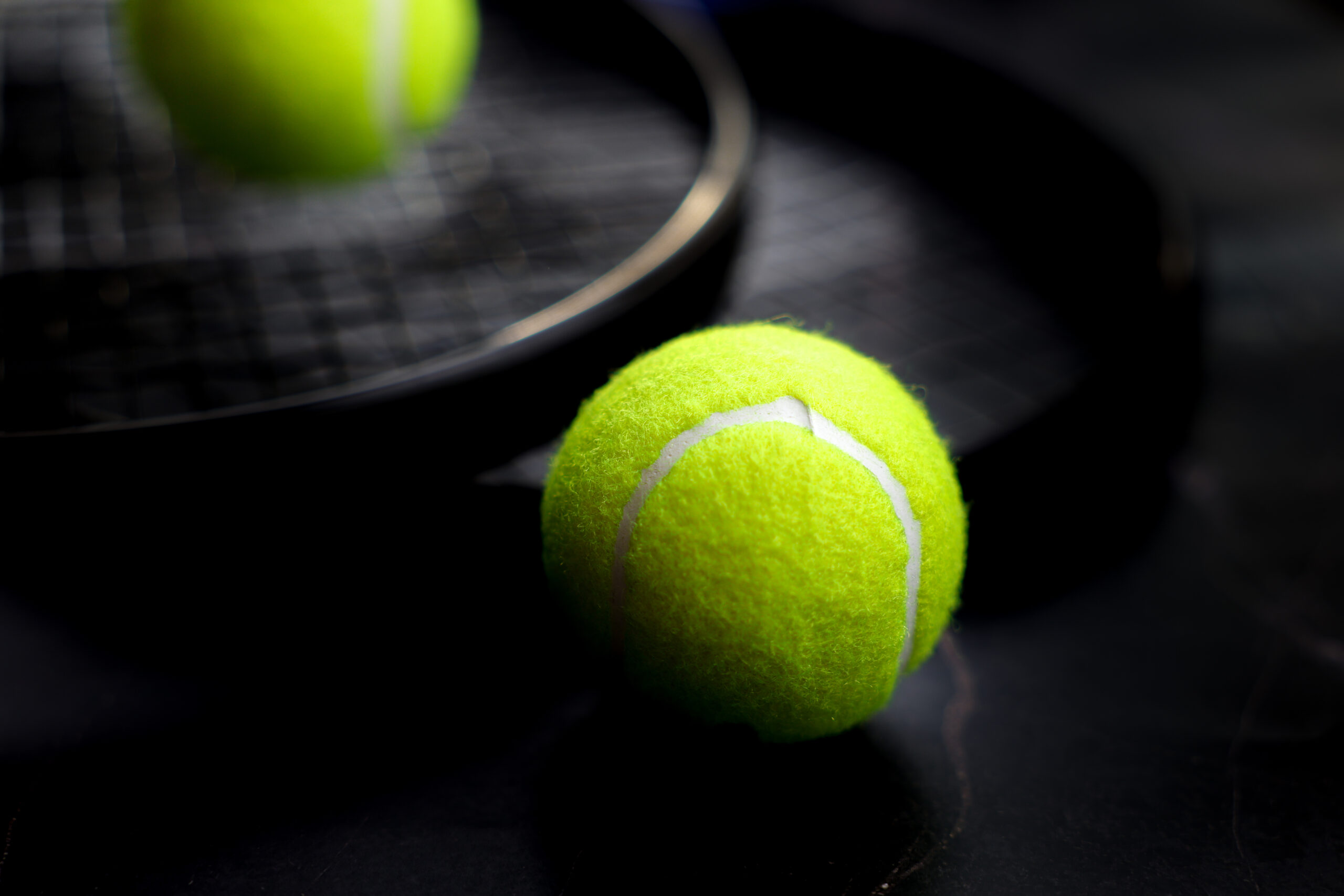 In the foreground lies a yellow tennis ball, behind it tennis rackets and a ball on a dark background are visible in out-of-focus. The concept of sports and competitions.